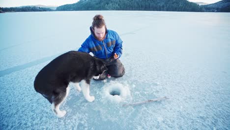 Caucasian-Man-Is-Fishing-Through-A-Hole-In-Ice-With-His-Alaskan-Malamute-Dog-At-Frozen-River-Near-Trondheim,-Norway