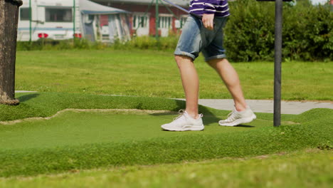 Young-guy-in-shorts-waiting-and-analyzing-court-before-hitting-miniature-golf-ball-putting-mini-golf-at-camping-having-fun-summer-games-party-activity-friends-at-golf-club-vacation-having-ice-cream