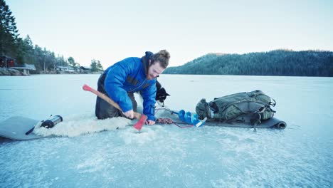 Un-Tipo-Pescando-En-Invierno-Rompiendo-El-Hielo-Del-Agujero-De-Un-Río-Congelado-Usando-Un-Hacha-En-Noruega