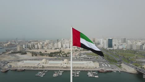 UAE-FLAG:-Drone-camera-circling-around-the-flag-of-United-Arab-Emirates-waving-in-the-air-at-Sharjah's-Flag-Island,-Sharjah-city-in-the-background