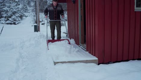 Schöner-Mann-In-Winterkleidung,-Der-Schnee-Mit-Einer-Schlittenschaufel-An-Der-Tür-Reinigt-Und-Drinnen-Hält