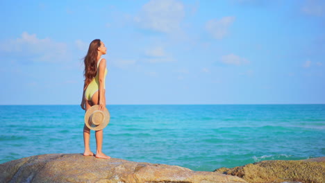 Standing-on-a-large-rock-a-pretty-woman-in-a-bathing-suit-and-sun-hat-in-hand-looks-over-the-ocean-waves