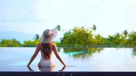 Back-view-of-woman-with-hat-sitting-on-infinity-pool-edge-looking-at-panorama