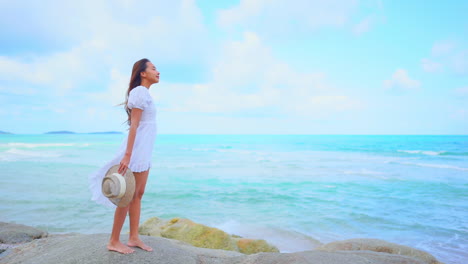 Standing-alone-on-a-large-rock,-a-pretty-young-woman-looks-out-to-sea