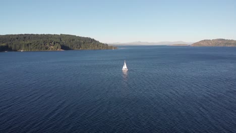 Distant-white-sailboat-on-deep-blue-lake-in-Patagonia,-tracking-aerial