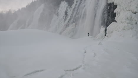 Girl-Standing-In-Thick-Icy-Surface-Near-A-Waterfall-In-Quebec-In-Winter---pan-up-wide-shot