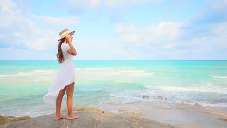 A-young-woman-in-a-sundress-holds-on-to-her-sun-hat-as-she-overlooks-the-incoming-waves