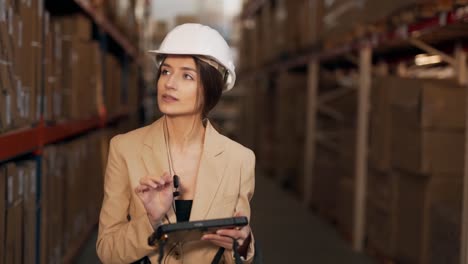 Beautiful-young-woman-in-a-business-suit-with-a-helmet-on-her-head-working-with-a-tablet-on-the-background-of-the-warehouse-with-boxes