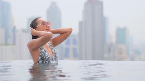 A-passionate-young-Asian-woman-touches-her-wet-hair-being-inside-infinity-swimming-pool-water-on-the-rooftop-of-a-luxury-hotel-with-city-skyscrapers-in-the-background