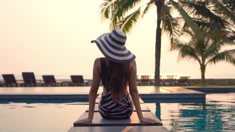 Sexy-woman-sitting-on-the-edge-of-the-swimming-pool-at-an-exotic-hotel-in-Florida-wearing-botched-spotty-monokini-and-striped-hat-looking-at-sunset-beach,-back-view-slow-motion-handheld