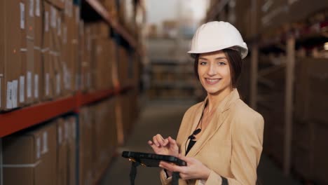 Portrait-of-a-beautiful-young-woman-in-a-business-suit-with-a-helmet-on-his-head-working-with-a-tablet-in-his-hands-on-the-background-of-the-warehouse-with-boxes