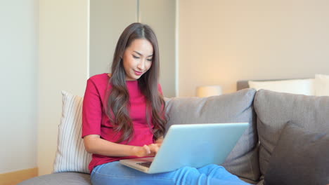 Asian-woman-sitting-on-the-sofa-with-her-laptop-on-laps-and-typing-on-keyboard-showing-the-beautiful-smile