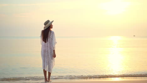 Rückansicht-Einer-Barfüßigen-Frau-Mit-Hut-Und-Weiß-Gekleidet-Am-Strand-Bei-Sonnenuntergang