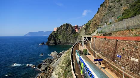 Static-view-of-stunning-train-station,-railway-and-tracks-alongside-beautiful-rocky-Italian-coastline-and-clear-blue-ocean-water-while-train-drives-through-on-sunny-summer-day