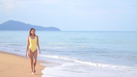Lonely-slim-asian-woman-in-swimsuit-walking-on-sandy-beach-by-tropical-sea-waves-on-sunny-day