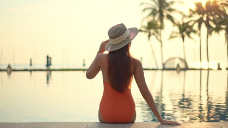 Back-view-of-Fit-female-model-wearing-an-orange-monokini-swimwear-suit-and-hat-sits-on-the-edge-of-the-swimming-pool-in-Turkey-at-sunset