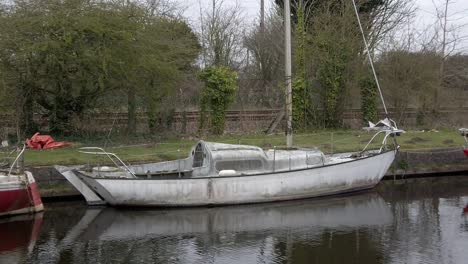 Small-dirty-sailboat-moored-on-narrow-rural-countryside-canal-marina