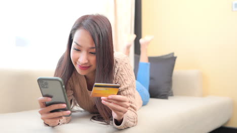 Junges-Lächelndes-Asiatisches-Mädchen,-Das-Auf-Sofa-Liegt-Und-Kreditkarte-Und-Smartphone-Hält