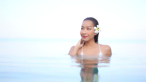 High-key-shot-of-beautiful-young-smiling-Asian-woman-with-flower-in-hair-relaxing-in-infinity-pool