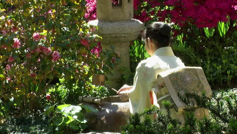 Eine-Japanische-Frau-Verwendet-Eine-Bambuskelle-Vor-Einer-Steinlaterne-In-Einem-Japanischen-Garten