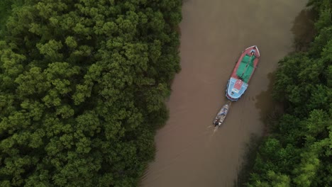 Cinematic-drone-top-down-shot-of-ship-carrying-small-boat-on-amazon-river-surrounded-by-green-rainforest-trees-during-sunset