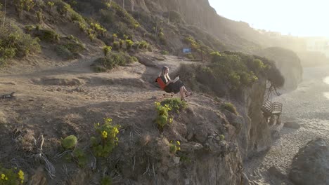 Aerial-View-Of-Beautiful-Young-Woman-Reading-On-Scenic-Cliff-At-Sunset