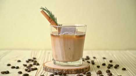 Milch-In-Schwarzes-Kaffeeglas-Mit-Eiswürfel,-Zimt-Und-Rosmarin-Auf-Holzhintergrund-Gießen