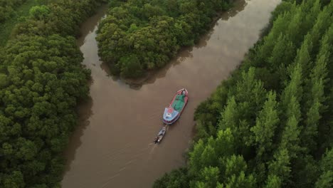 Cinematic-drone-orbit-shot-of-ship-carrying-small-boat-on-amazon-river-surrounded-by-green-rainforest-trees-during-sunset