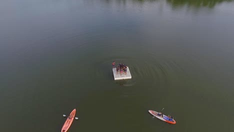Drone-view-of-a-group-of-children-jumping-from-a-summer-camp-lake-platform,-and-two-kayaks-surrounding-it