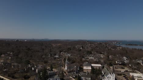 A-long,-slow-descending-shot-of-Boston,-MA-USA-in-the-distance,-revealing-the-scenic-downtown-area-of-Hingham,-MA-in-the-foreground