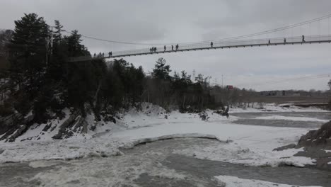 People-Walking-Across-Suspension-Bridge-Over-Flowing-Icy-River-At-Chutes-De-La-Chaudiere-In-Levis,-Quebec-City,-Canada