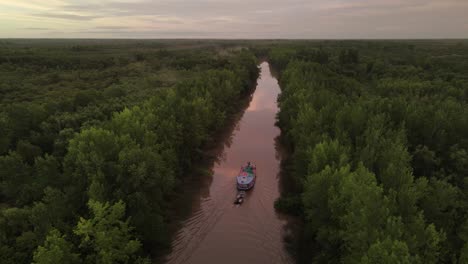 Cinematic-drone-shot-of-ship-carrying-small-boat-on-amazon-river-surrounded-by-green-rainforest-trees-during-sunset