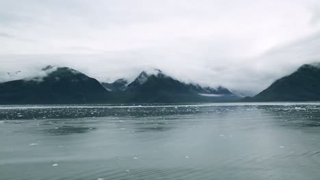 Calm-Ocean-Horizon-with-Small-Glacier-Ice-Pieces-Floating-and-Mountains-in-Background-on-a-Dark-Gloomy-Overcast-Cloudy-Day,-Alaska-USA