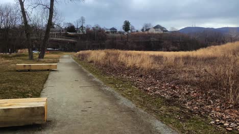A-walking-path-with-benches-in-a-public-green-space-next-to-a-small-city