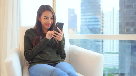 Hübsche-Asiatische-Frau,-Die-Zu-Hause-Smartphone-Verwendet,-Stadtbildhintergrund
