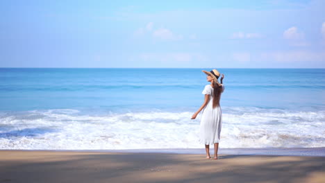 Asian-Woman-standing-on-the-beach-by-the-sea-water-when-big-foamy-tides-roll-on-sandy-beach-in-summer-wearing-a-white-sundress-and-straw-hat---slow-motion-static-shot
