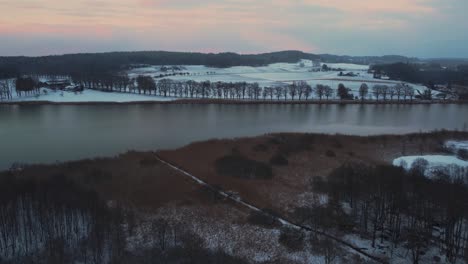 4K-UHD-drone-aerial-of-a-natural-lake-surrounded-by-hills-and-fields-and-an-oak-tree-avenue-in-Bavaria,-Germany,-in-winter-with-snow-during-a-red-cloud-sky-sunset