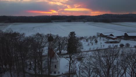 Eine-Alte-Kapellenkirche-Mit-Einer-Runden-Kuppel-Auf-Einem-Hügel,-Umgeben-Von-Bäumen-Im-Winter-In-Bayern,-Deutschland,-Mit-Schnee-Auf-Den-Feldern-Von-Oben-Gesehen,-Luftdrohnenaufnahmen-Während-Des-Sonnenuntergangs-Mit-Roten-Wolken-Und-Himmel