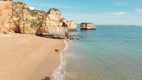 Fly-over-golden-sands-beach-approaching-jagged-limestone-rock-formations,-Lagos,-Algarve