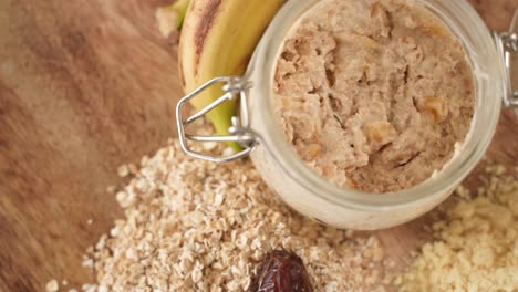 Oatmeal-Porridge-in-Jar-next-to-Bananas,-Dried-Dates-on-Wooden-Background