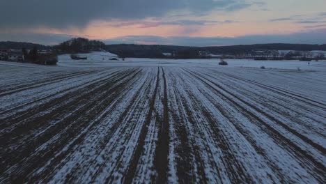 A-rural-field-surrounded-by-trees-in-winter-in-Bavaria,-Germany,-with-snow-on-the-ground-seen-from-above,-aerial-drone-footage-while-sunset-with-red-clouds-and-sky