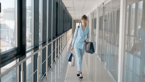 Stylish-young-girl-with-packages-walks-down-the-glass-corridor-and-smiles