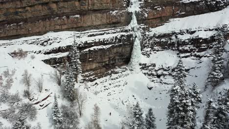 Aerial-View-of-Lonely-Person-Walking-on-Snow-Under-Cliffs-With-Frozen-Waterfall-on-Cold-Winter-Day,-Tilt-Up-Drone-Shot