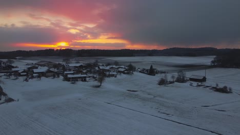 A-rural-village-surrounded-by-trees-in-winter-in-Bavaria,-Germany,-with-snow-on-the-fields-and-roofs-seen-from-above,-aerial-drone-footage-while-sunset-with-red-clouds-and-sky