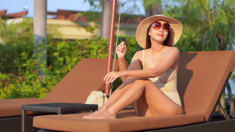 Adorable-luxury-Asian-lady-suntanning-on-lying-on-the-deck-chair-at-tropical-Thailand-resort-wearing-beige-monokini,-sunglasses,-and-straw-hat