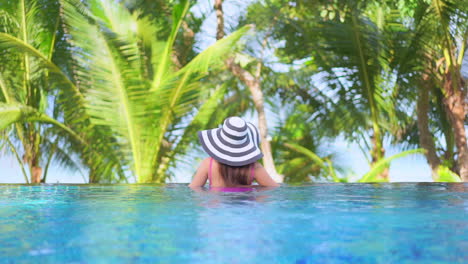 Back-view-of-woman-with-large-hat-and-pink-swimsuit-in-pool-edge