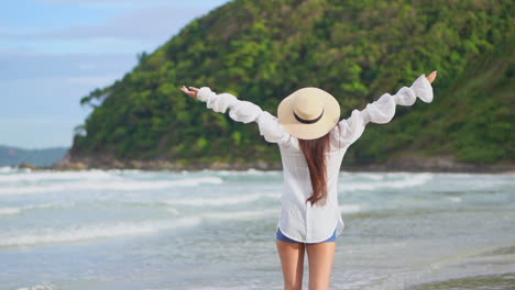 Back-view-of-Asian-girl-wearing-a-white-blouse-and-shorts-spreading-and-raising-her-arms-during-a-morning-walk-on-island's-beach,-sea-tides-and-green-mountains-on-background,-Slow-motion