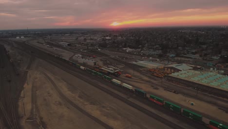 Zooming-out-cinematic-top-city-aerial-view-of-downtown-Roseville,-California-with-industrial-buildings,-shops-and-cargo-train-passing-down-the-railroad-with-a-vivid-orange-pink-sunset-in-background
