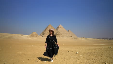 Girl-In-Black-Dress-Walking-In-The-Desert,-Pyramids-In-The-Background-In-Cairo,-Egypt---wide-shot