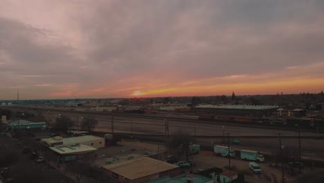 Bottom-up-cinematic-top-city-aerial-view-of-downtown-Roseville,-California-with-industrial-buildings,-shops-and-cargo-train-passing-down-the-railroad-with-a-vivid-orange-pink-sunset-in-background